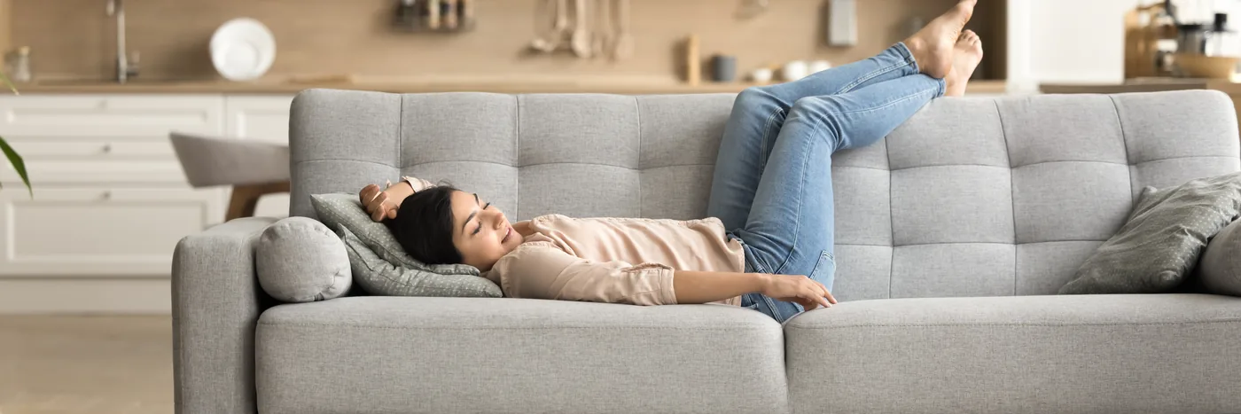 A woman lying back along a grey sofa with her eyes closed and her legs stretched out over the arm.