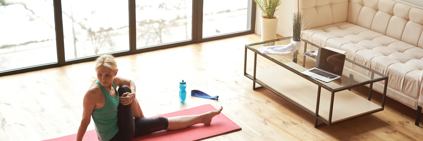 A woman stretching on a pink exercise mat on the floor of a living room, a laptop open on the coffee table beside her.