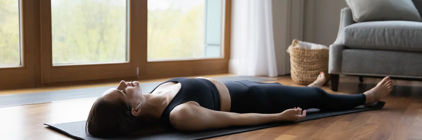 A woman lying flat on her back on a dark exercise mat on a wooden floor, arms at her sides, in a bright room with tall windows.