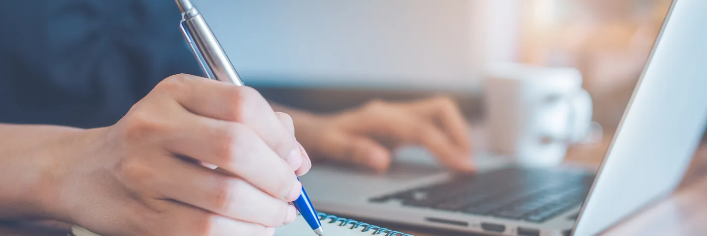 A hand writing notes on a small pad beside an open laptop and a mug of coffee.